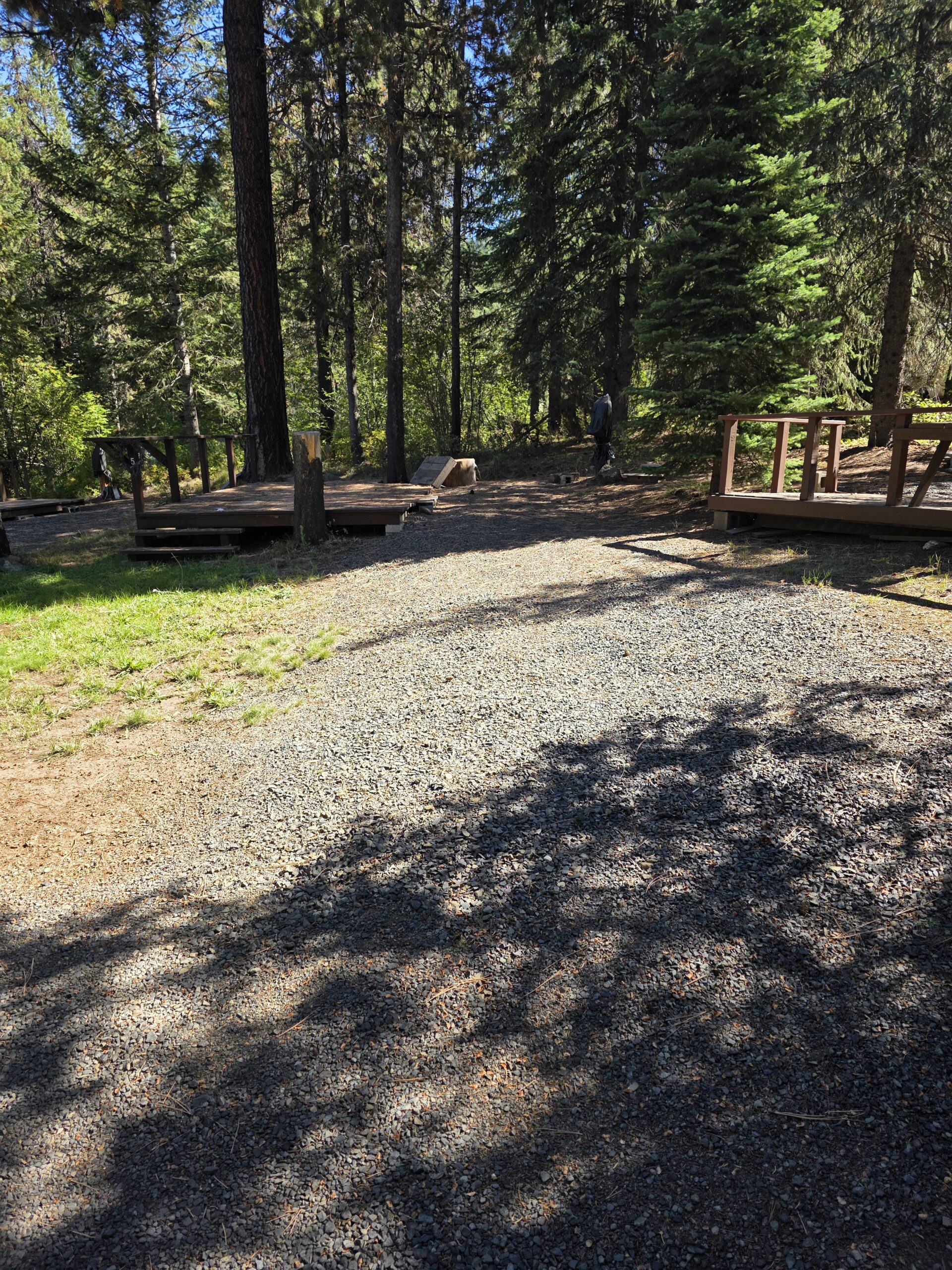 A gravel path leads through a wooded area with tall evergreen trees, bordered by wooden decks on both sides; a person stands in the distance among the trees.