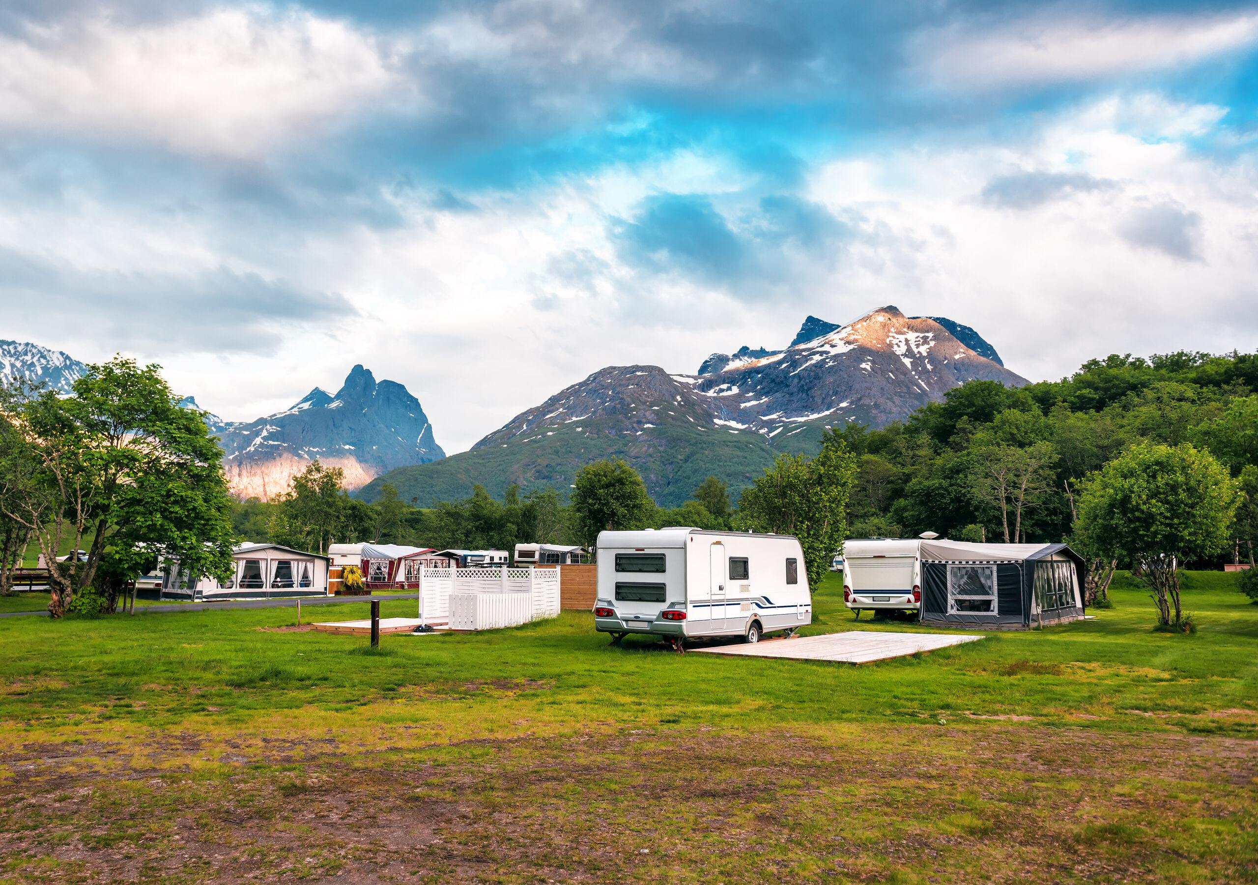 Caravans and tents are set up on a grassy campground surrounded by trees, with tall, snow-capped mountains and a partly cloudy sky in the background.